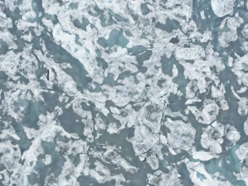 Aerial view of a glacier featuring abstract patterns of white ice and grey water in a natural environment, taken with an overhead angle.