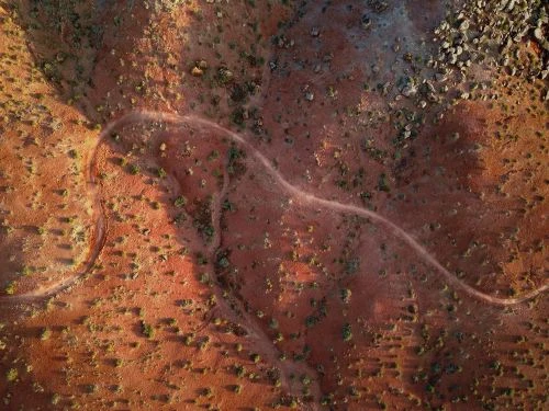 Aerial view of a winding dirt path through a red desert landscape with sparse green vegetation and rocky areas, captured from above.