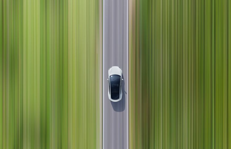 Aerial view of a white electric car driving along a grey asphalt road, flanked by blurred, green grass, creating a sense of speed.