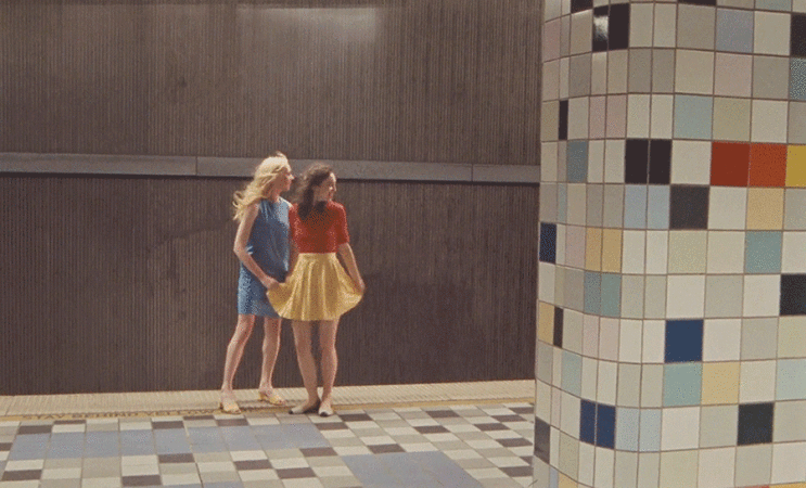 Two young women in dresses pose at subway station, one in blue and one in yellow skirt and red top, tiled walls and floors.