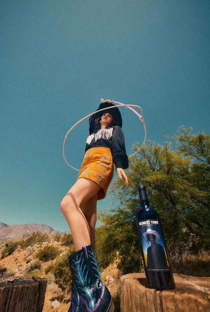 Woman in cowboy boots with a lasso and a wine bottle featuring a portrait, in a desert environment against a blue sky backdrop.