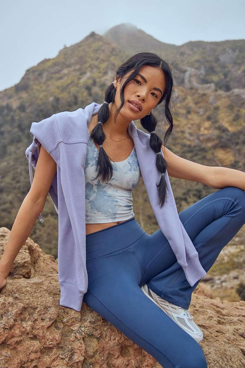 Athletic woman with braided hair sits on a rock, wearing a lilac-colored hoodie, blue top, and leggings, with a mountain background.
