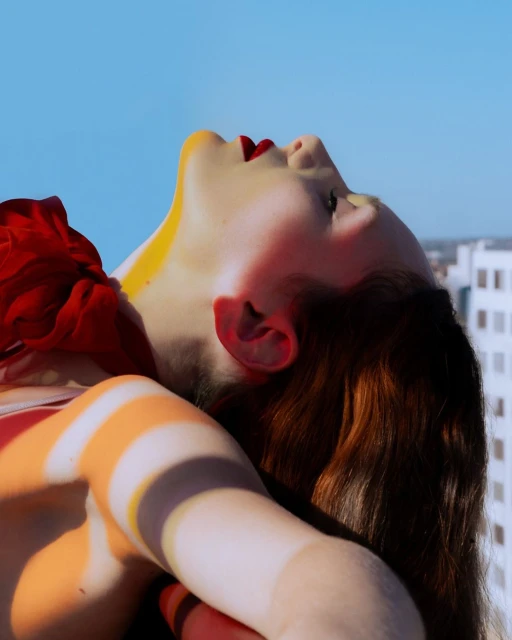A woman with red hair and red lipstick lies on a red surface against a blue sky, with a red scarf and orange lighting.