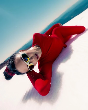 Model in a bright red jumpsuit and white sunglasses lying on white sand dunes against a blue sky