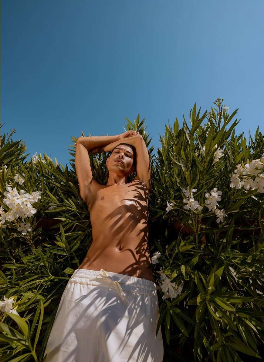 Low angle view of a shirtless adult male lying amongst green foliage and white flowers against a bright blue sky in outdoor light.