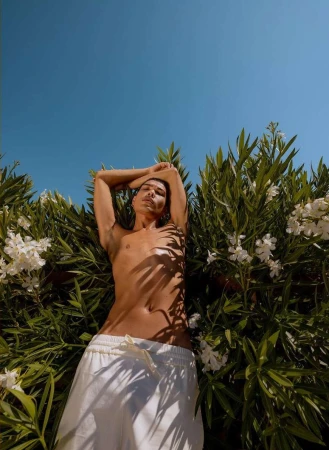 Low angle view of a shirtless adult male lying amongst green foliage and white flowers against a bright blue sky in outdoor light.