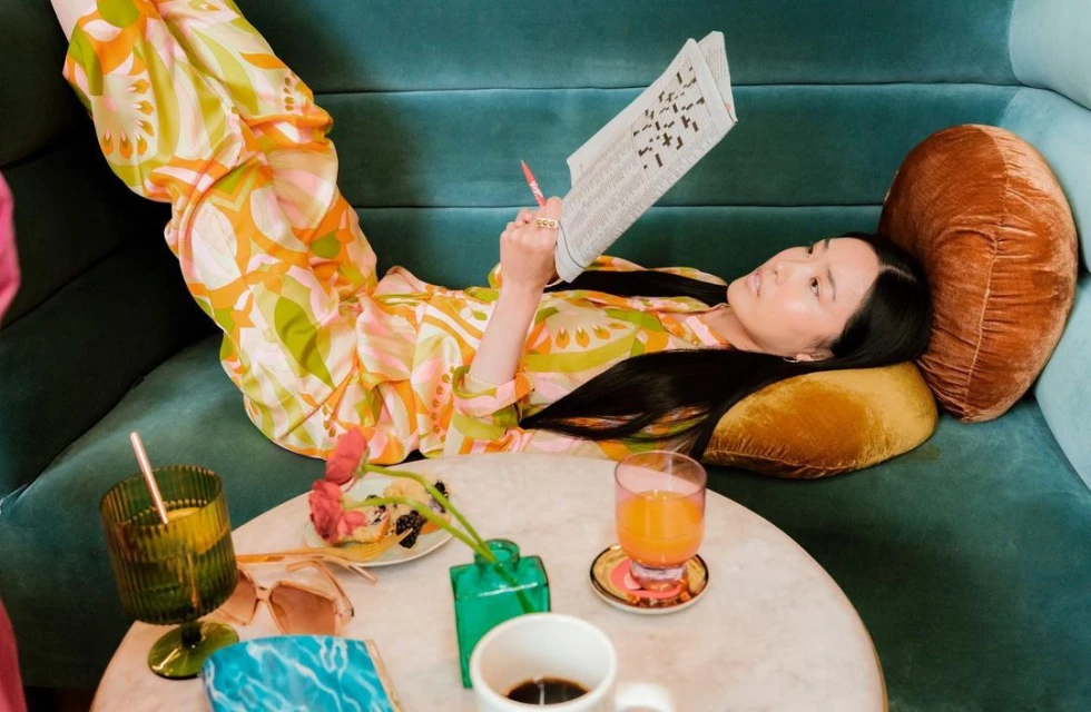 Woman in patterned pajamas lies on a teal couch, doing a crossword puzzle with drinks and snacks on a marble table nearby.