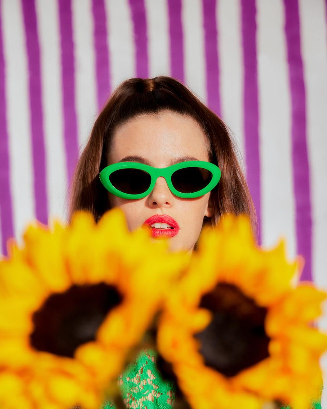 Woman in green sunglasses poses with sunflowers against purple and white striped background in a fashion studio portrait.