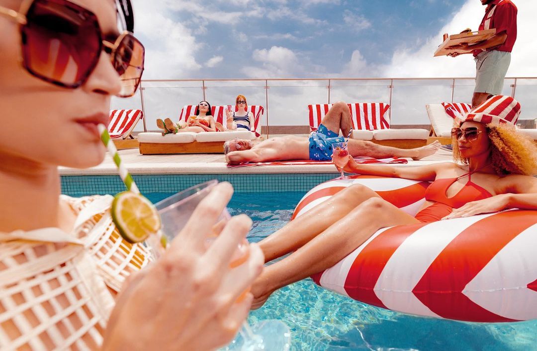 People relaxing by the pool on lounge chairs with cocktails, attended by a server with food, on a rooftop under a sunny, partly cloudy sky.