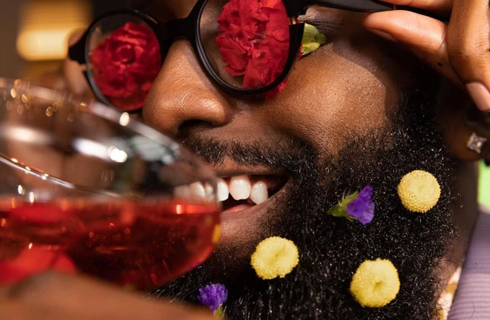 Close-up of an adult Black man with a beard decorated with flowers drinking red wine and wearing black-rimmed glasses with floral reflections.