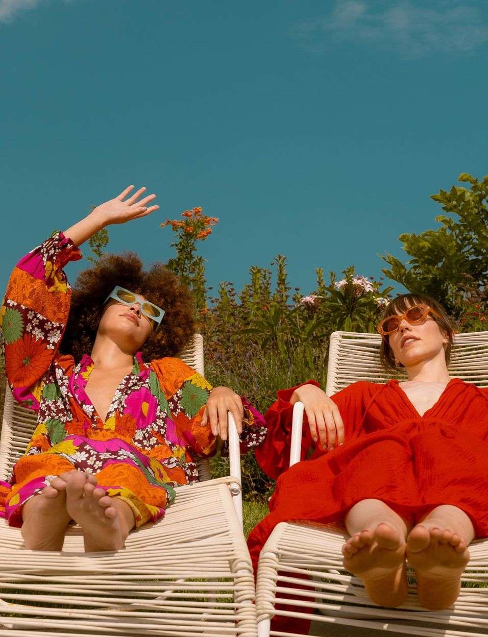 Two young women in vibrant dresses recline in chairs on a sunny day, showcasing a blend of fashion and leisure in an outdoor setting.