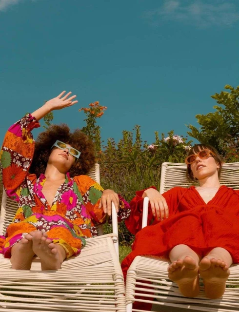Two young women in vibrant dresses recline in chairs on a sunny day, showcasing a blend of fashion and leisure in an outdoor setting.