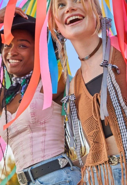 Two smiling women, one with a hat, stand against a bright blue sky, colorful ribbons surround them, creating a joyful, summery scene.
