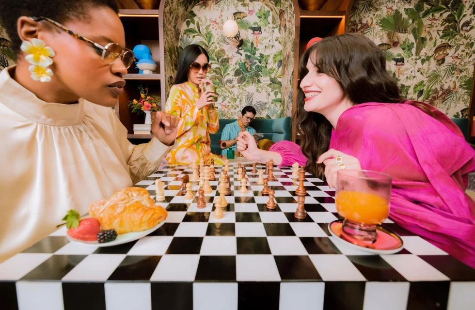 Four people gather around a black-and-white checkered table in an eclectic interior with tropical wallpaper, drinks, and a chessboard.