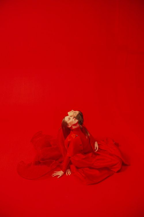 Two women in matching red dresses pose against a vivid red backdrop, their necks arched back in a synchronized dance or fashion display.