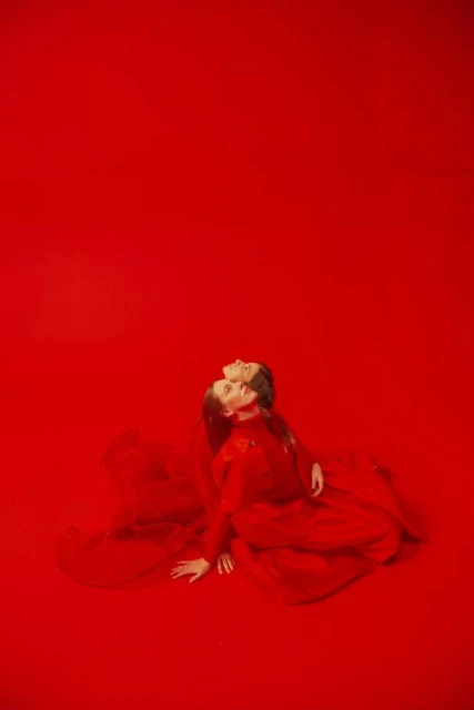 Two women in matching red dresses pose against a vivid red backdrop, their necks arched back in a synchronized dance or fashion display.