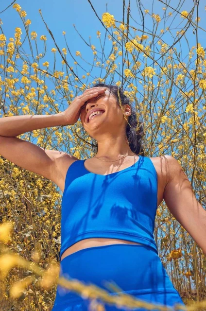 A smiling woman in blue athletic wear shields her eyes in an upward shot, surrounded by yellow flowers against a clear sky.