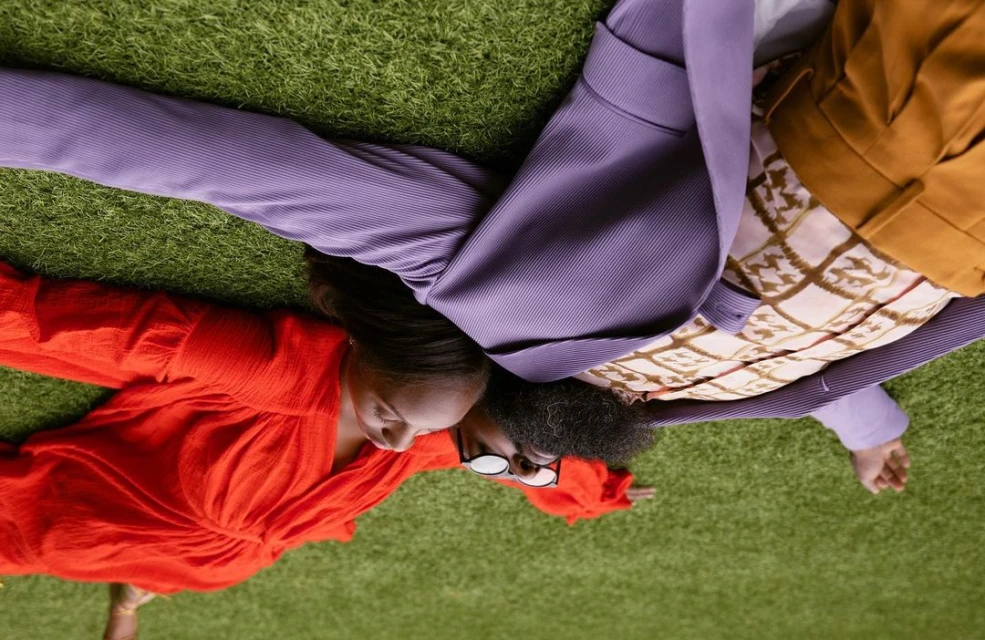 Two adults lie head-to-head on green grass; an adult female wears a red dress, while an adult male sports a purple jacket and glasses.