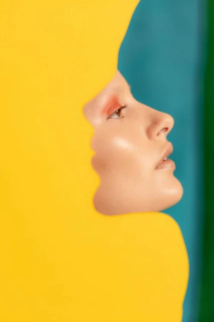 Profile of woman's face revealed in a bright yellow shape against a backdrop of teal, green and dark blue color blocks. Beauty shot.