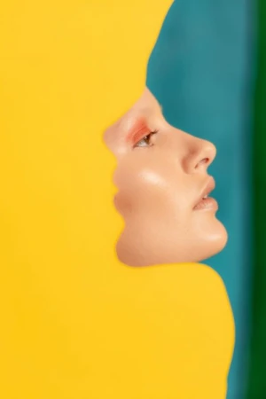Profile of woman's face revealed in a bright yellow shape against a backdrop of teal, green and dark blue color blocks. Beauty shot.
