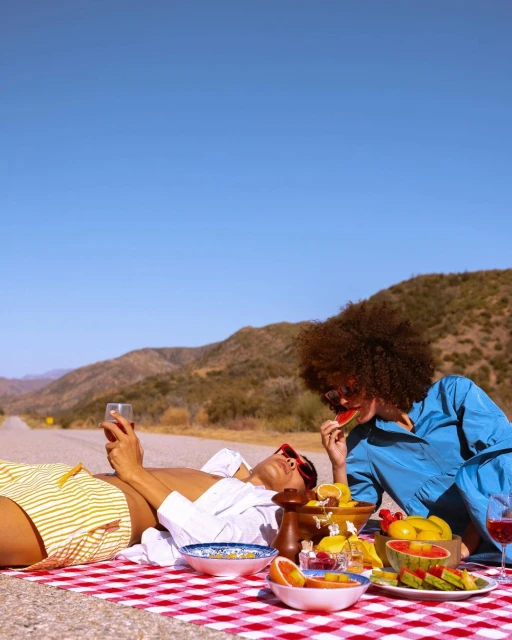 Two women enjoy a roadside picnic with watermelon, citrus fruit, and wine on a red and white checkered blanket under a clear blue sky.