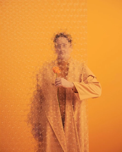 Man in a tan robe holding an orange flower stands in front of an orange background, partially obscured by patterned plastic wrap.