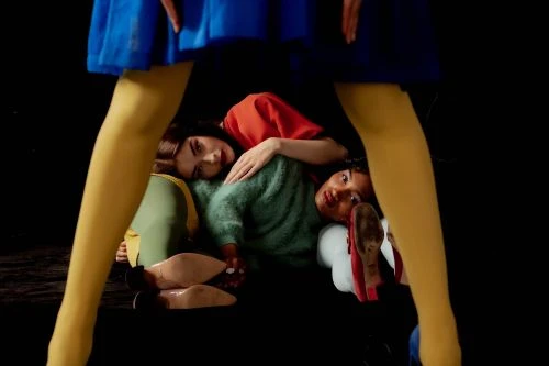High-angle shot of two women in sweaters posed in a pile under a dress, showcasing vibrant colors and an intimate, artistic composition.