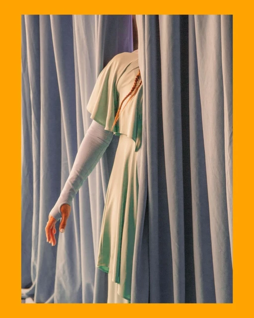 Elegant arm extends from behind blue drapes. Woman wears a flowing pale green dress. Braided hair detail. Studio lighting.