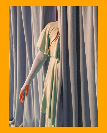 Elegant arm extends from behind blue drapes. Woman wears a flowing pale green dress. Braided hair detail. Studio lighting.