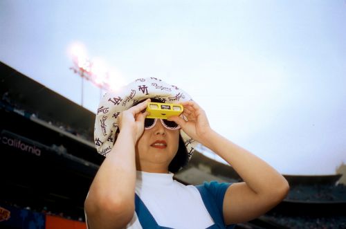 Woman wearing a Los Angeles Dodgers hat and solar eclipse glasses at an outdoor baseball stadium on a sunny day.