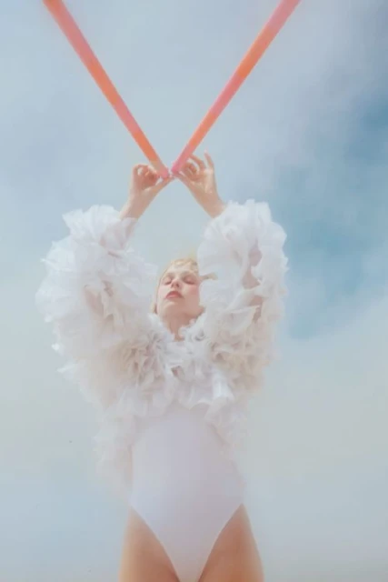 Artistic portrait of a woman in a white bodysuit and feathered bolero, holding two coral-colored ribbons against a pastel sky background.
