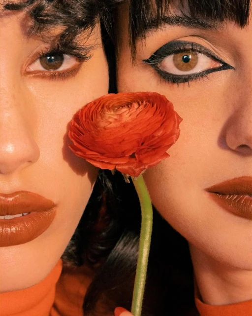 Close-up of two adult women with matching red-orange lipstick, separated by a vibrant coral ranunculus flower in a beauty shot.