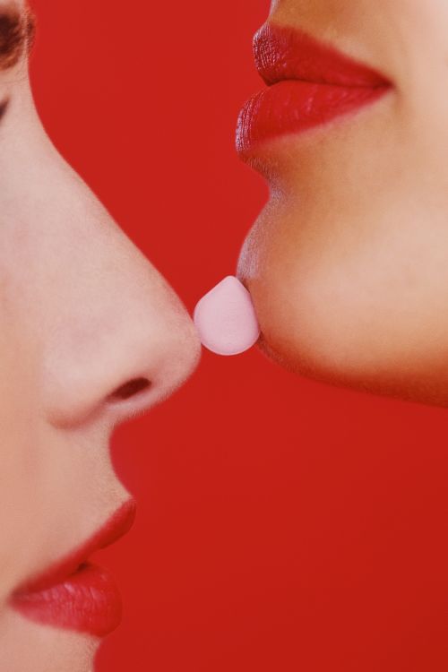 Close-up profile shot of two women facing each other with a pink pill held between their noses, against a solid red background