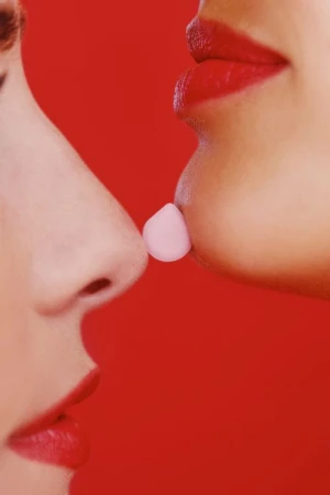 Close-up profile shot of two women facing each other with a pink pill held between their noses, against a solid red background