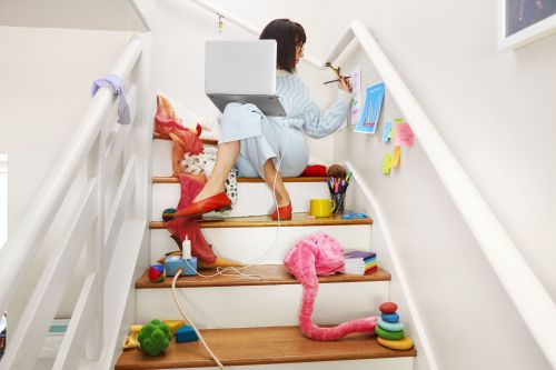 Woman sitting on stairs with laptop painting with color brush, surrounded by children's toys, books, and art supplies.