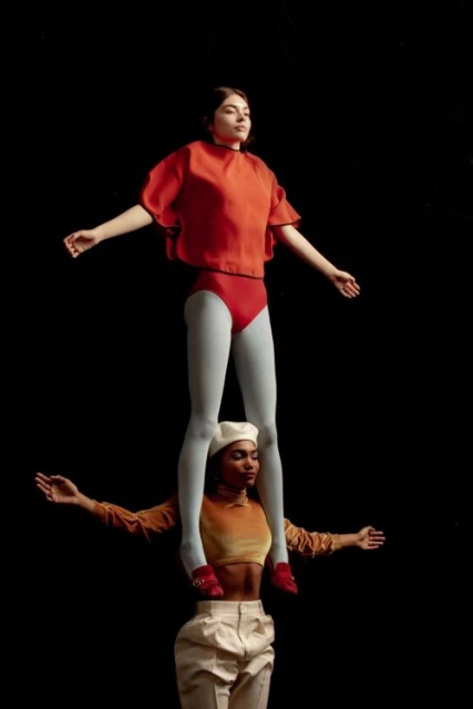 Two female acrobats strike a pose against a dark background. One woman in orange stands atop the other, who wears a beret and white pants.