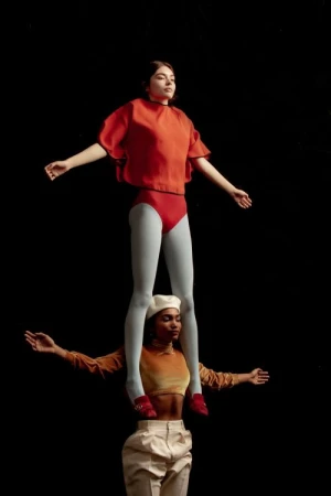 Two female acrobats strike a pose against a dark background. One woman in orange stands atop the other, who wears a beret and white pants.