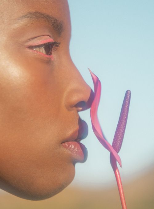 Profile of Black woman with pink eye makeup and flower near her face against a blue sky in a beauty or fashion shot.