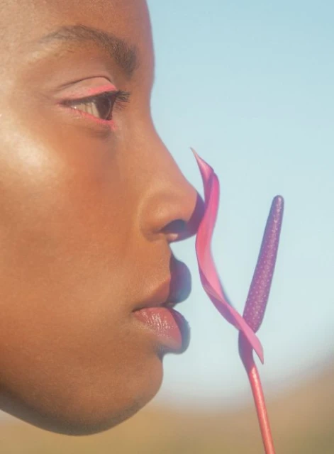 Profile of Black woman with pink eye makeup and flower near her face against a blue sky in a beauty or fashion shot.