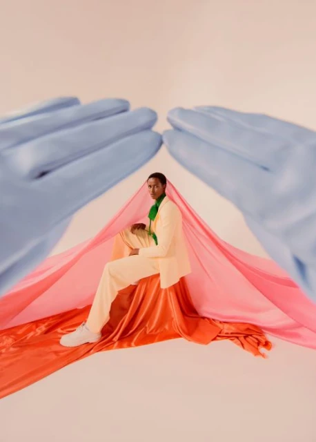Person in light-yellow suit sits on draped fabric with medical gloves overhead, creating a surreal fashion portrait in a studio.