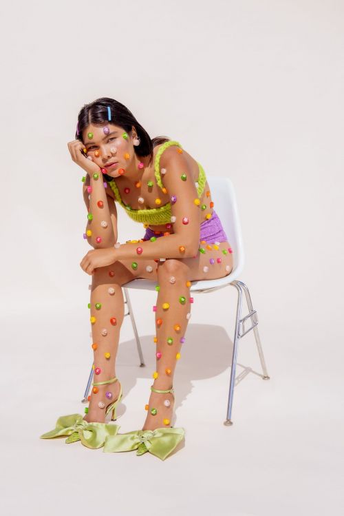 Woman sits on a white chair covered in colorful candy dots. She wears a knit top, shorts, and matching high heels with oversized bows.