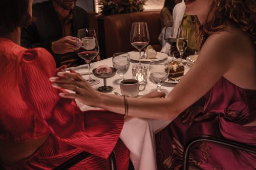 A dimly lit restaurant scene featuring a table setting with wine glasses, dessert plates, and several people enjoying a meal in an indoor setting.