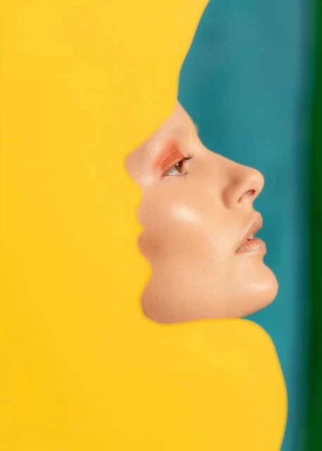 Profile of a fair-skinned woman with coral eye shadow, framed by graphic yellow and teal shapes in a modern beauty shot.