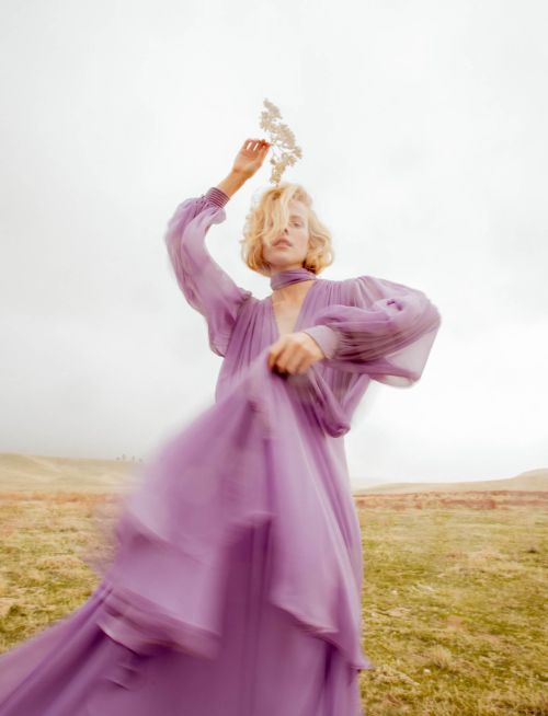 Woman in a flowing lavender dress and bare feet posing in a field with a sprig of small white flowers held above her head.