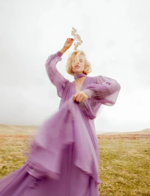Woman in a flowing lavender dress and bare feet posing in a field with a sprig of small white flowers held above her head.