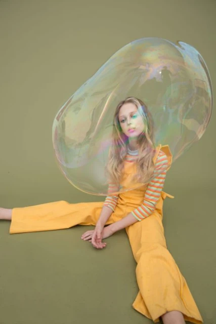 Young woman in a yellow jumpsuit sits indoors with a large soap bubble around her head in a studio setting, creating an eye-catching portrait.