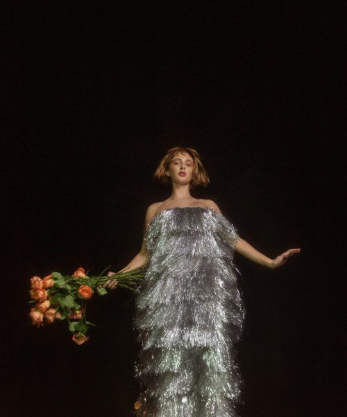 Woman in a silver fringe dress holds a bouquet of roses against a dark backdrop, creating a striking contrast in a studio setting.