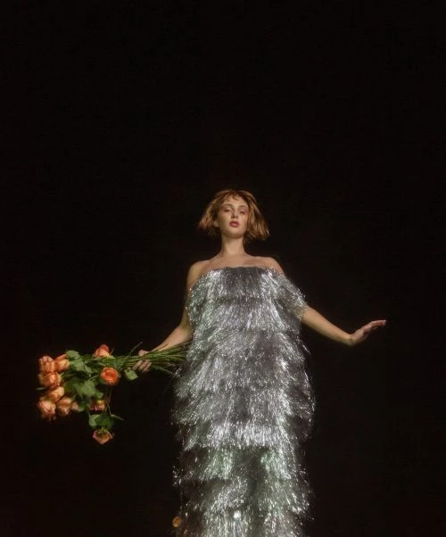 Woman in a silver fringe dress holds a bouquet of roses against a dark backdrop, creating a striking contrast in a studio setting.