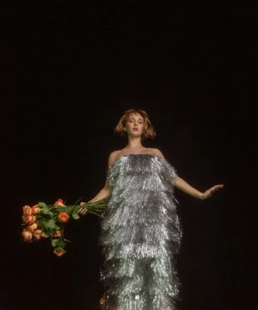 Woman in a silver fringe dress holds a bouquet of roses against a dark backdrop, creating a striking contrast in a studio setting.