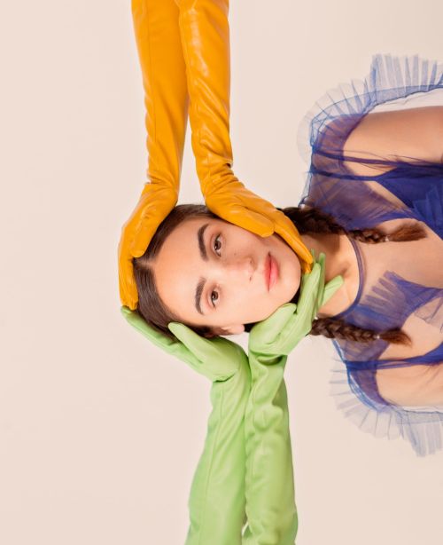 Eye-level studio portrait of a young woman with braided hair, wearing a blue dress, and styled with long yellow and green gloves.